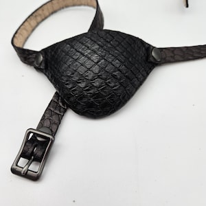 May include: A black leather eye patch with a textured snakeskin pattern. The patch is attached to a black leather strap with a metal buckle.
