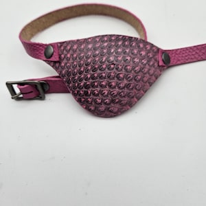Op de afbeelding: Een roze leren ooglapje met een getextureerd, schubbig ontwerp. Het ooglapje is bevestigd aan een roze leren band met een gesp.