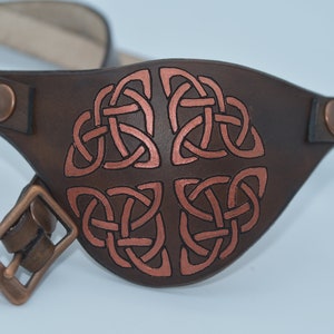 Op de afbeelding: Een bruin leren ooglapje met een koperkleurig Keltisch knoopontwerp. Het ooglapje heeft een band met een gesp en twee koperkleurige drukknopen.