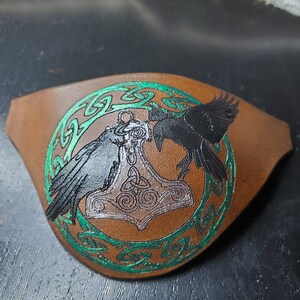 May include: A brown leather eye patch with a green Celtic knot design. The center of the patch features a silver Mjolnir hammer with two black ravens on either side.