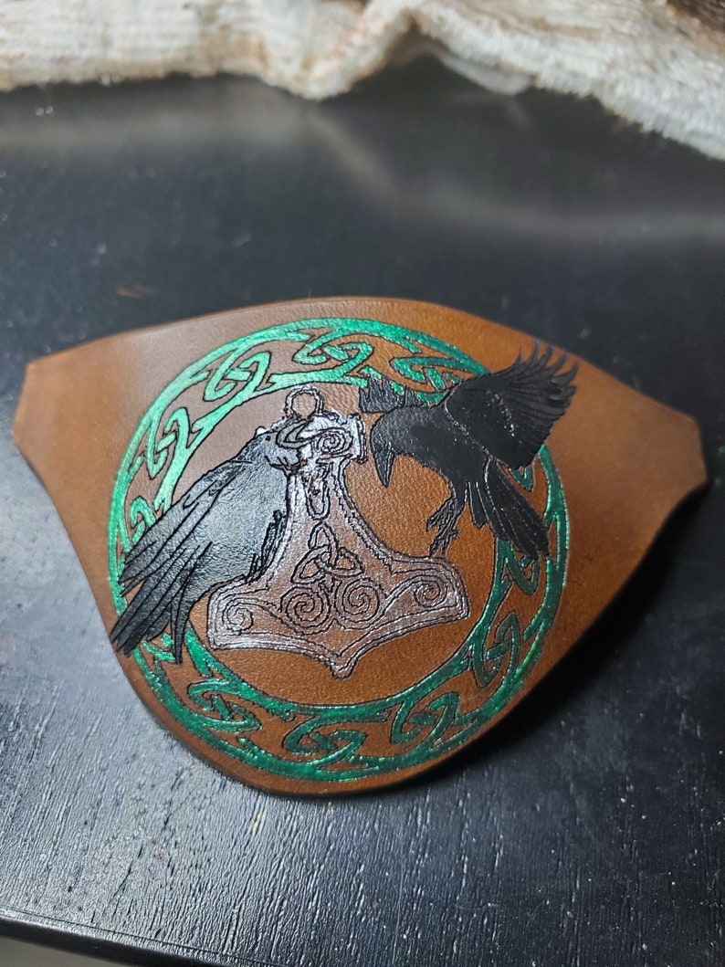 May include: A brown leather eye patch with a green Celtic knot design. The center of the patch features a silver Mjolnir hammer with two black ravens on either side.