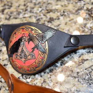 May include: Black leather eye patch with a red, gold, and black design featuring two ravens and a hammer. The design is surrounded by a gold Celtic knot.