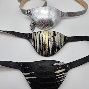 May include: Three leather eye patches with adjustable straps. The eye patches are black, silver, and black with gold and silver stripes.