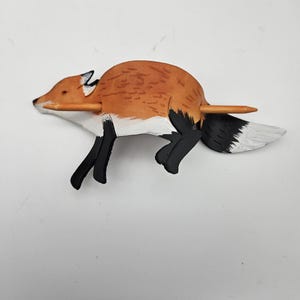 Op de afbeelding: Een houten vossenbeeldje met een potlood erdoorheen. De vos is beschilderd met oranje, zwarte, witte en grijze details. De staart van de vos is wit met grijze punten. Het potlood is lichtbruin.