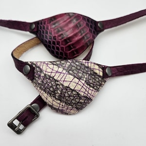 May include: Two leather eye patches with adjustable straps. One patch is a deep purple with a reptile skin pattern, and the other is off-white with a black and purple grid design. Both have dark purple straps and metal hardware.