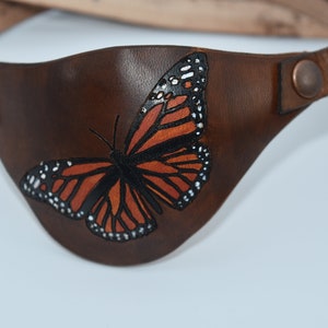 May include: A brown leather eye patch with a painted monarch butterfly design. The butterfly has orange, black, and white wings.