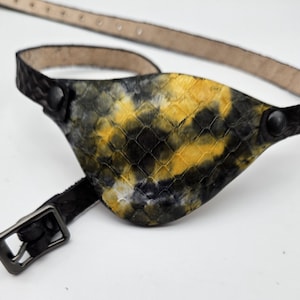 May include: A black leather eye patch with a yellow and black tie-dye pattern. The patch has a buckle closure and a strap.