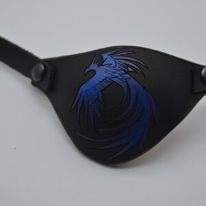 Puede incluir: Un parche para el ojo de cuero negro con un dise&ntilde;o de f&eacute;nix azul y morado.