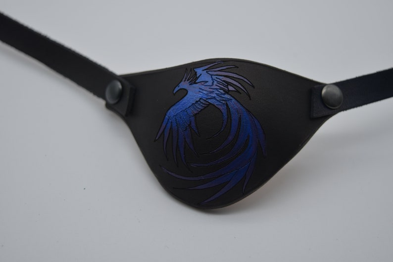 Puede incluir: Un parche para el ojo de cuero negro con un dise&ntilde;o de f&eacute;nix azul y morado.