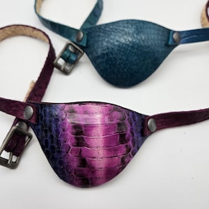 May include: Two leather eye patches. One is teal blue and the other is a purple and black snakeskin pattern.