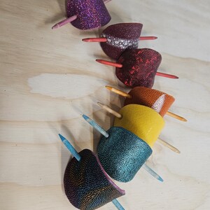 May include: Six colorful hair clips with wooden sticks. The clips are made of leather and have a textured surface. The colors are purple, red, orange, yellow, blue, and green.