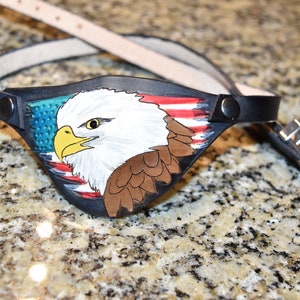 May include: A black leather eye patch with an American flag and a bald eagle painted on it. The eye patch has a strap that can be adjusted to fit different head sizes.