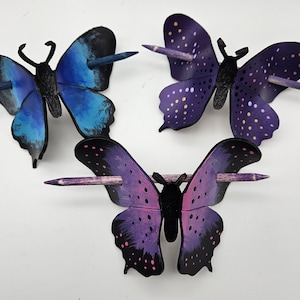 Op de afbeelding: Drie decoratieve haarspelden in de vorm van vlinders. Eén vlinder is blauw, de tweede is paars met gele stippen en de derde is paars met zwarte vlekken. Elke vlinder heeft een houten speld.