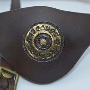 K&ouml;nnte beinhalten: Ein braunes Leder-Augenpflaster mit einem goldfarbenen Design einer 12 Gauge-Schrotflinte.