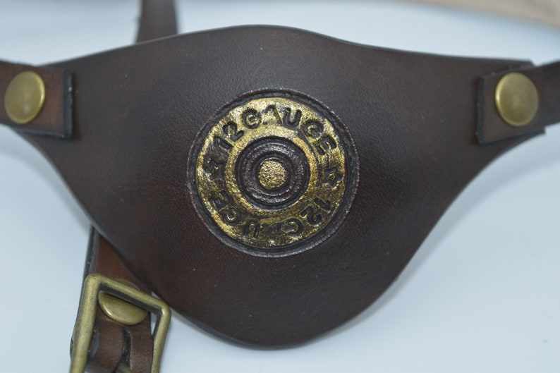 K&ouml;nnte beinhalten: Ein braunes Leder-Augenpflaster mit einem goldfarbenen Design einer 12 Gauge-Schrotflinte.