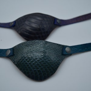 May include: Two leather eye patches with adjustable straps. The top patch is dark purple with a textured surface. The bottom patch is dark green with a textured surface.
