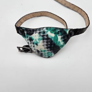 May include: A black leather eye patch with a green and white snakeskin pattern. The patch has a buckle fastening and is attached to a black leather strap.