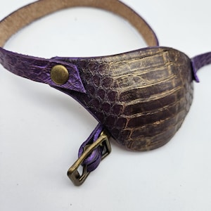 May include: A purple leather eye patch with a gold embossed crocodile pattern and a brass buckle.