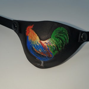 Op de afbeelding: Een zwart leren ooglapje met een kleurrijke haan erop geschilderd. De haan is blauw, groen, rood, oranje en geel.