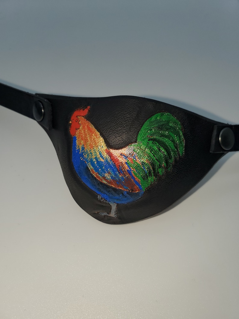 Op de afbeelding: Een zwart leren ooglapje met een kleurrijke haan erop geschilderd. De haan is blauw, groen, rood, oranje en geel.