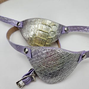 May include: Two eye patches with a metallic silver and textured design. The patches have a lavender strap and buckle. The eye patches are designed to be worn over one eye.