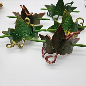 May include: Several decorative leaf-shaped hair pins. The leaves are primarily green and brown with gold and copper accents. Each leaf is attached to a green stick-like pin, designed to secure the hair. The pins are arranged on a white surface.