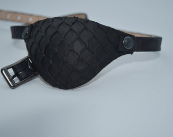 Könnte beinhalten: Ein schwarzes Leder-Augenpflaster mit einem Schuppenmuster und einem Schnallenverschluss. Das Pflaster ist an einem schwarzen Lederband befestigt.