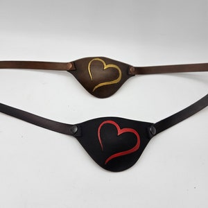 Può includere: Due parches per occhi in pelle con cinturini regolabili. Un parches per occhi è marrone con un design a forma di cuore dorato, l'altro è nero con un design a forma di cuore rosso.