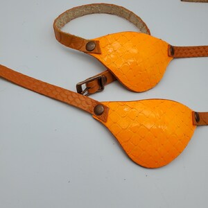 May include: Two orange leather eye patches with brown leather straps and metal buckles. The eye patches have a textured surface.
