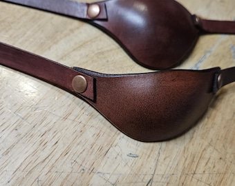 Könnte beinhalten: Zwei braune Leder-Augenklappen mit verstellbaren Riemen und kupferfarbenen Metallschnallen.