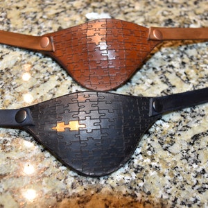 Può includere: Due parches per occhi in pelle con un design a puzzle. Uno è in pelle marrone e l'altro in pelle nera.