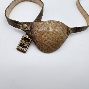 May include: A brown leather eye patch with a buckle and strap. The eye patch has a textured, scaly pattern.