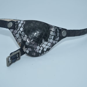 May include: A black leather eye patch with a silver snakeskin pattern. The patch is attached to a black leather strap with a silver buckle.