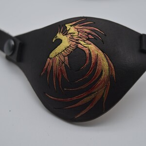 Puede incluir: Un parche para el ojo de cuero negro con un dise&ntilde;o de f&eacute;nix dorado y rojo.