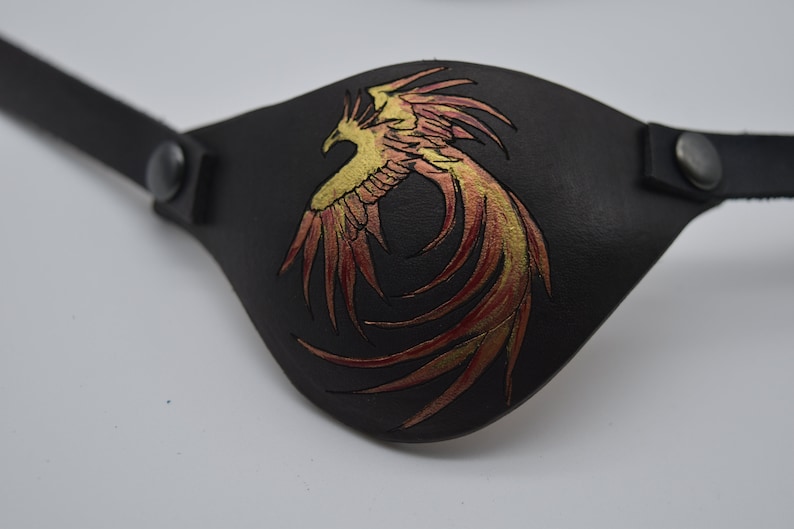 Puede incluir: Un parche para el ojo de cuero negro con un dise&ntilde;o de f&eacute;nix dorado y rojo.