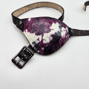 May include: A black leather eye patch with a purple and white tie-dye pattern. The patch has a buckle closure and is adjustable.