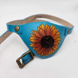 May include: A blue leather eye patch with a sunflower design. The patch has a strap and a buckle.