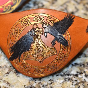 May include: A brown leather eye patch with a gold Celtic knot design and two black ravens flanking a gold Mjolnir hammer.