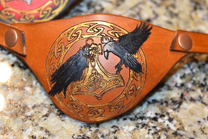 May include: A brown leather eye patch with a gold Celtic knot design and two black ravens flanking a gold Mjolnir hammer.