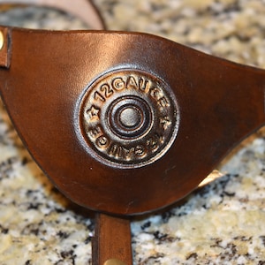 May include: A brown leather eye patch with a gold-colored 12 gauge shotgun shell casing embossed on the front.