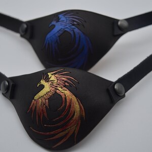 Puede incluir: Dos parches para ojos de cuero negro con correas ajustables. Un parche presenta un dise&ntilde;o de f&eacute;nix azul, el otro un dise&ntilde;o de f&eacute;nix dorado.
