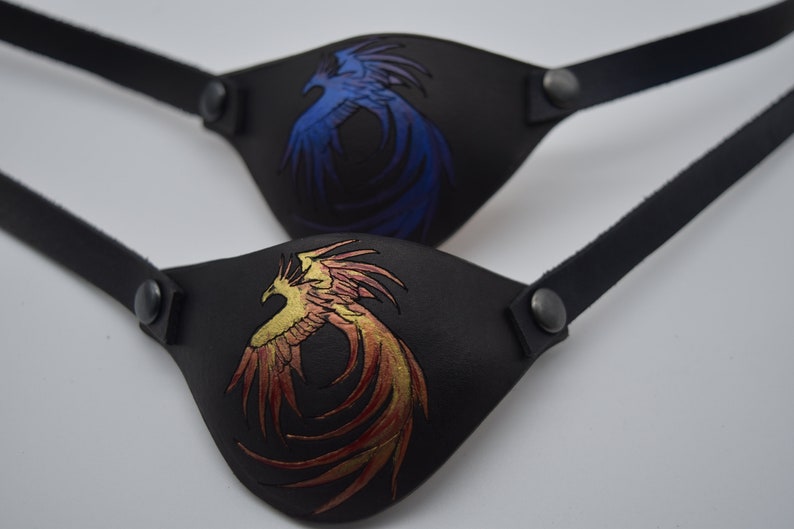 Puede incluir: Dos parches para ojos de cuero negro con correas ajustables. Un parche presenta un dise&ntilde;o de f&eacute;nix azul, el otro un dise&ntilde;o de f&eacute;nix dorado.