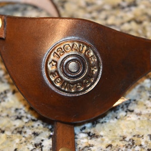 K&ouml;nnte beinhalten: Ein braunes Leder-Augenpflaster mit einer goldfarbenen 12 Gauge-Schrotpatronenh&uuml;lse, die auf der Vorderseite gepr&auml;gt ist.