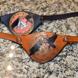 May include: Two leather eye patches with a black and brown strap. The eye patches feature a circle design with two black ravens and a silver hammer in the centre. One eye patch has a red background and the other has a brown background.