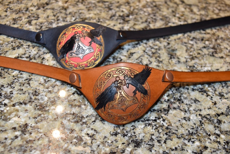 May include: Two leather eye patches with a black and brown strap. The eye patches feature a circle design with two black ravens and a silver hammer in the center. One eye patch has a red background and the other has a brown background.