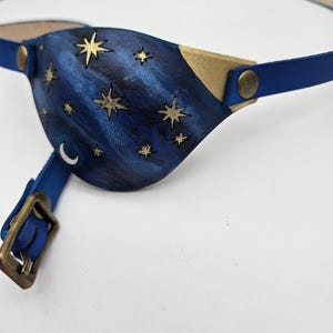 May include: A blue leather eye patch with a gold trim and a design of stars on a dark blue background.