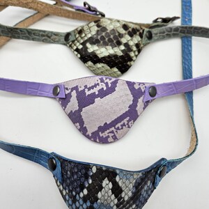 May include: Three handmade eye patches with straps. The top patch has a black and white snakeskin pattern. The middle patch is purple and white. The bottom patch is blue, black, and white. All patches have a curved shape and are made of leather.