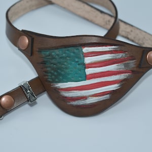 May include: A brown leather eye patch with a distressed American flag design. The patch is attached to a brown leather strap with copper colored buckles.