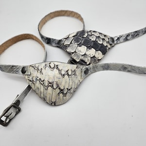May include: Two white leather eye patches with a snakeskin pattern. One patch has a black and white pattern, the other has a gray and white pattern. The patches are attached to leather straps with buckles.