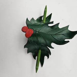 Op de afbeelding: Donkergroen leren haaraccessoire in de vorm van een hulstblad met drie rode bessen. Twee groene houten stokjes zijn door het blad gestoken. Het accessoire is ontworpen om het haar op zijn plaats te houden.